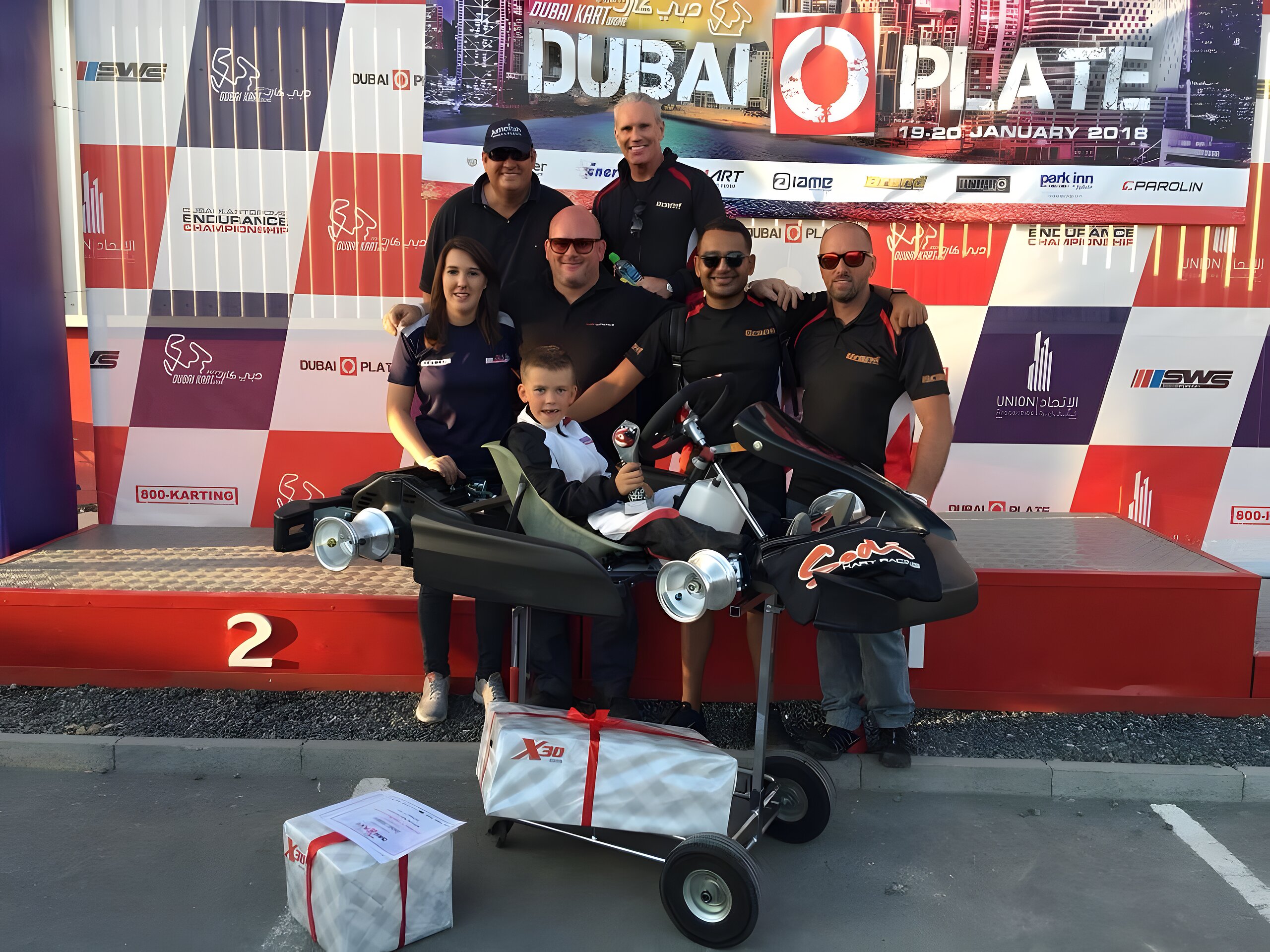 The whole crew around the kart on the Dubai O Plate podium, prizes stacked on the trolley